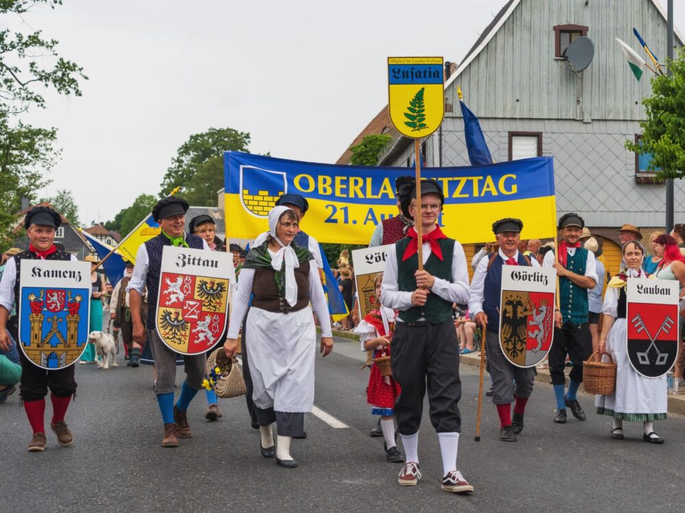 Menschen laufen in traditioneller Tracht mit verschiedenen Stadtwappen bei einem Umzug die Straße entlang.