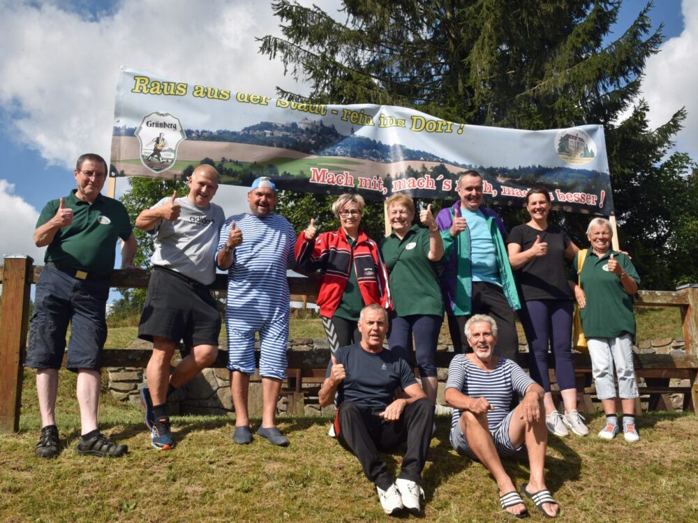 Eine Gruppe lächelt fröhlich in die Kamera und hält einen Banner mit der Aufschrift „Raus aus der Stadt – rein ins Dorf!“ in die Höhe.