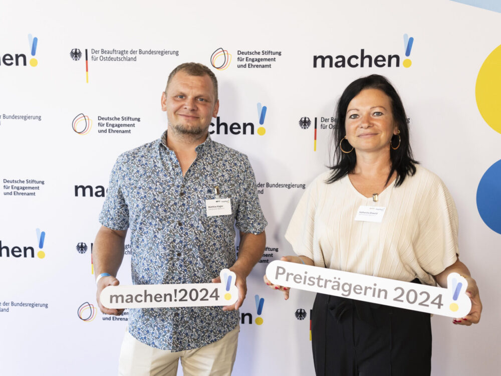 Ein Mann und eine Frau halten die Auszeichung der Preisverleihung von „machen!2024“ in die Kamera.