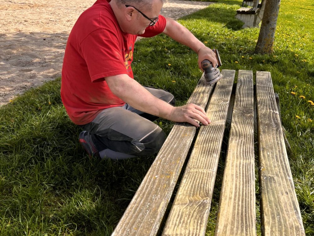 Ein Mann schleift die Holzbalken einer Sitzbank mit einem Handschleifer ab.