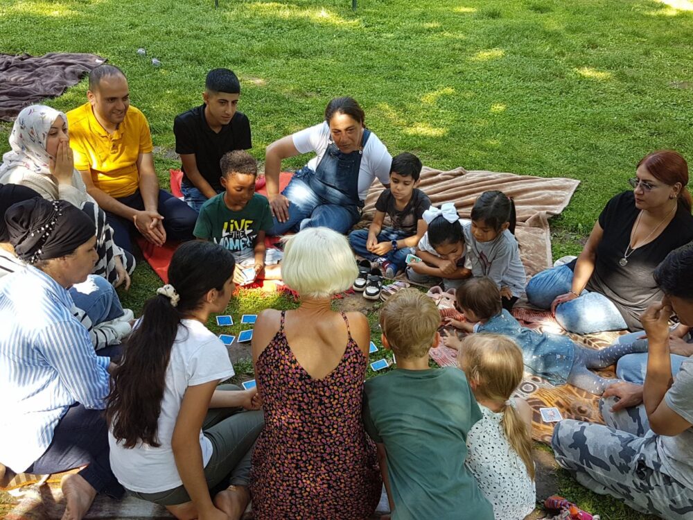 Geflüchtete Familien sitzen zusammen mit Ehrenamtlichen auf Decken im Park und spielen ein Kartenspiel.