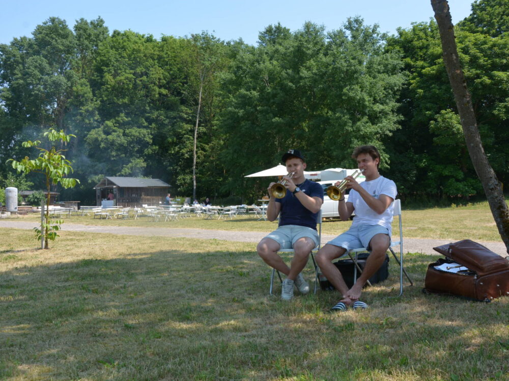 Zwei junge Männer sitzen auf Klappstühlen auf einer Wiese und spielen Trompete.