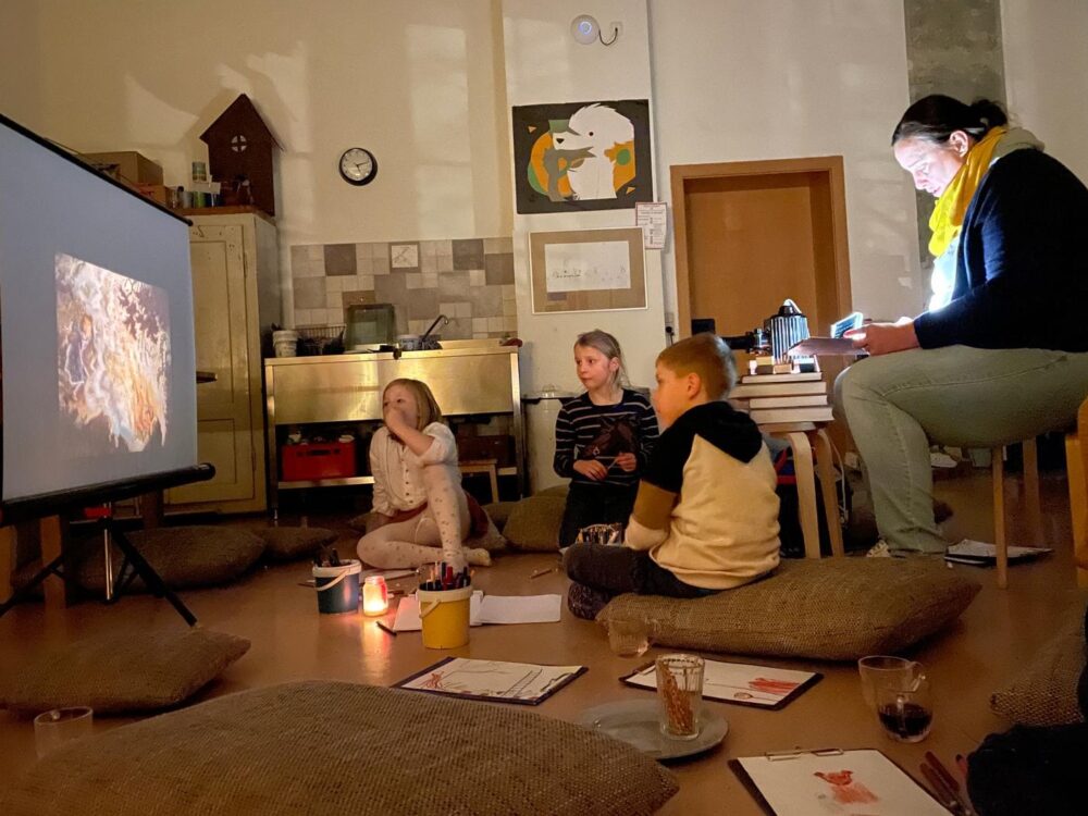 Drei Kinder sitzen mit Papier und Stiften auf dem Boden und schauen auf eine Projektion auf einer Leinwand.