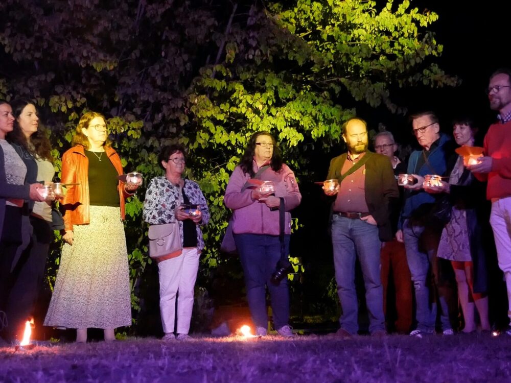 Eine Gruppe von Menschen steht im Dunkeln auf einer Wiese und hält Gläser mit brennenden Kerzen in den Händen.