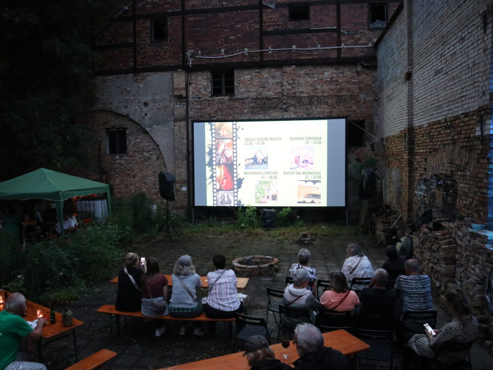 Menschen sitzen im Freien vor einer Leinwand.