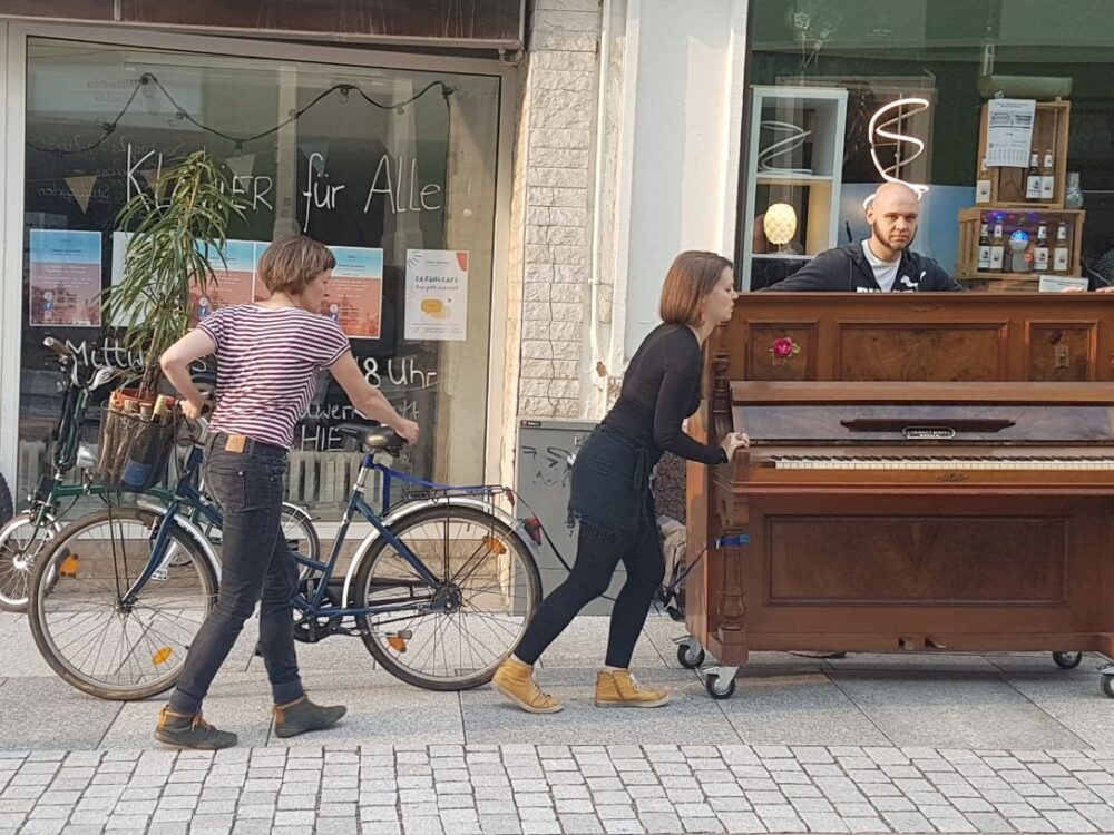 Zwei Frauen und ein Mann schieben ein Klavier auf Rollen über den Gehweg.