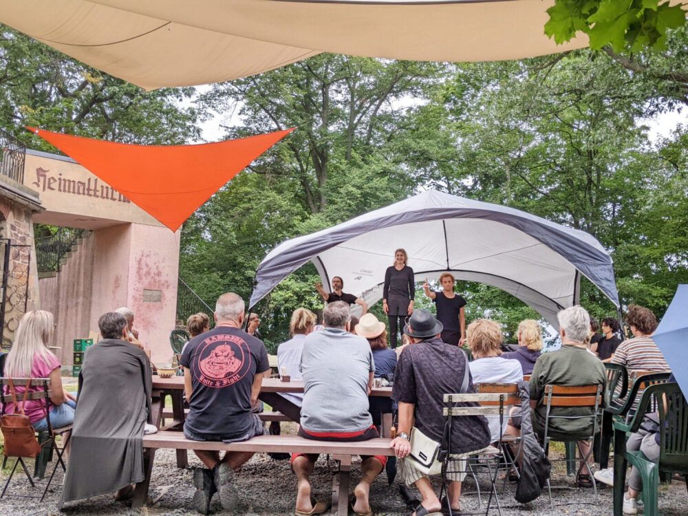 Zuschauende beobachten eine Theateraufführung auf einer kleinen Bühne unter freiem Himmel.