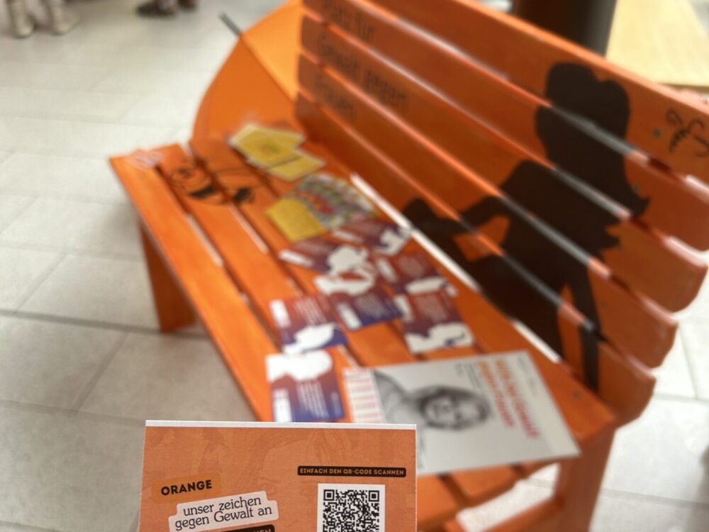 Auf einer orangen Holzbank liegen Informationsflyer. Im Vordergrund hält eine Hand einen Zettel mit einem QR-Code in die Kamera.