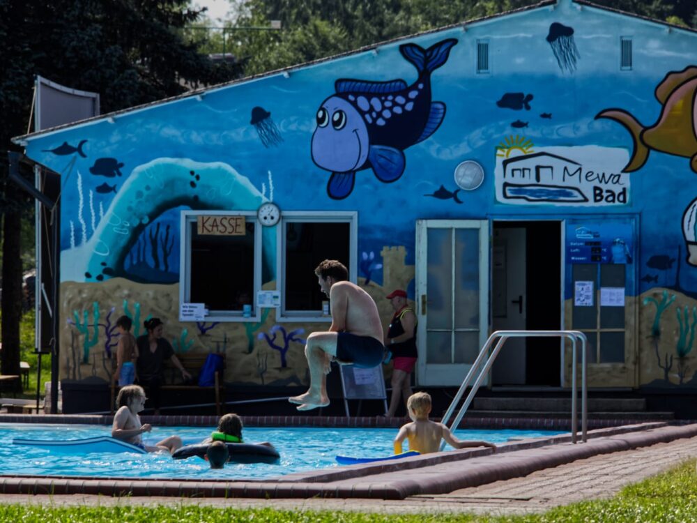 Ein Mann springt vom Beckenrand zu planschenden Kindern ins Schwimmbecken.