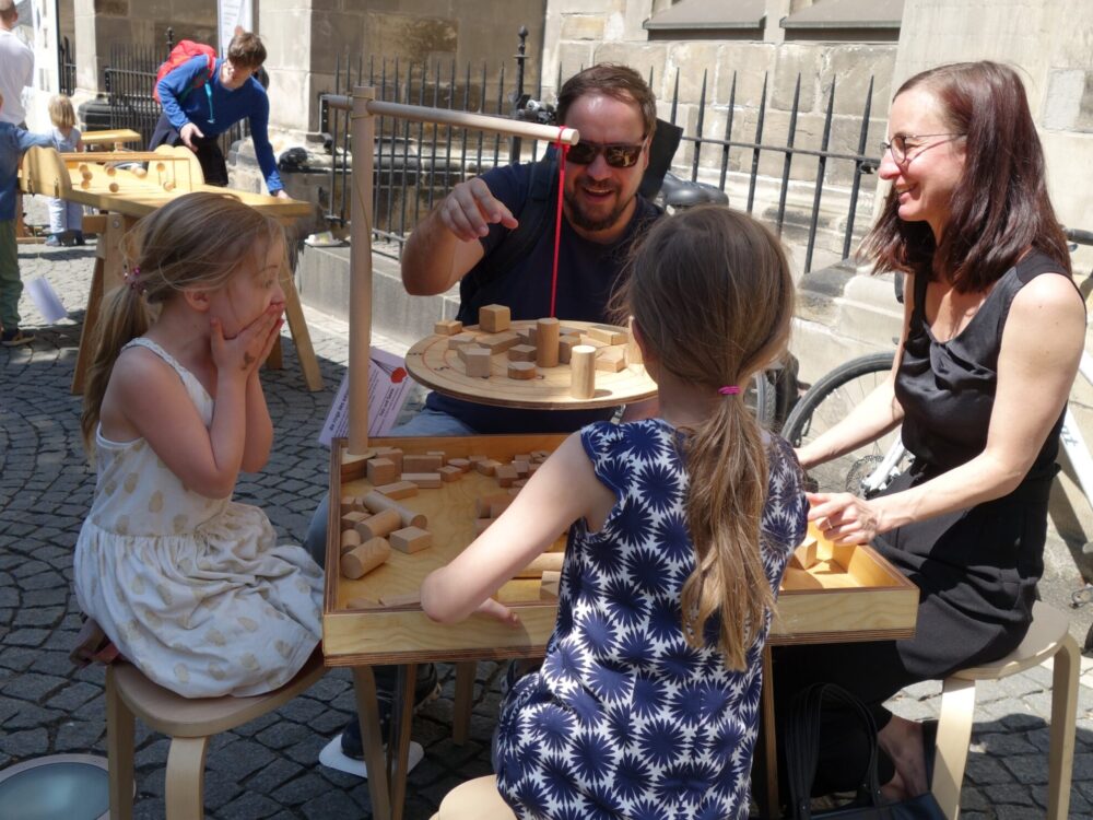 Zwei Mädchen spielen mit ihren Eltern an einem Holztisch auf einem Dorfplatz ein Geschicklichkeitsspiel.