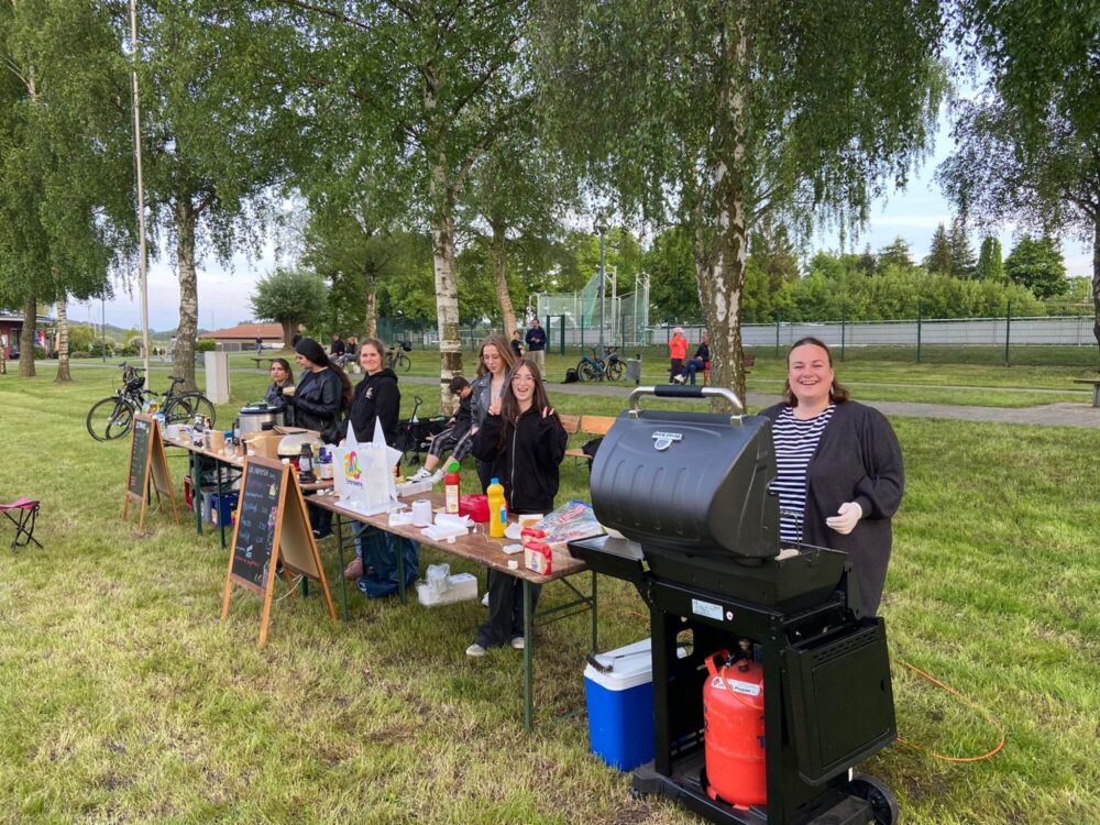 Eine Gruppe Jugendlicher steht hinter Biertischen und einem großen Grill um Essen zu verkaufen.