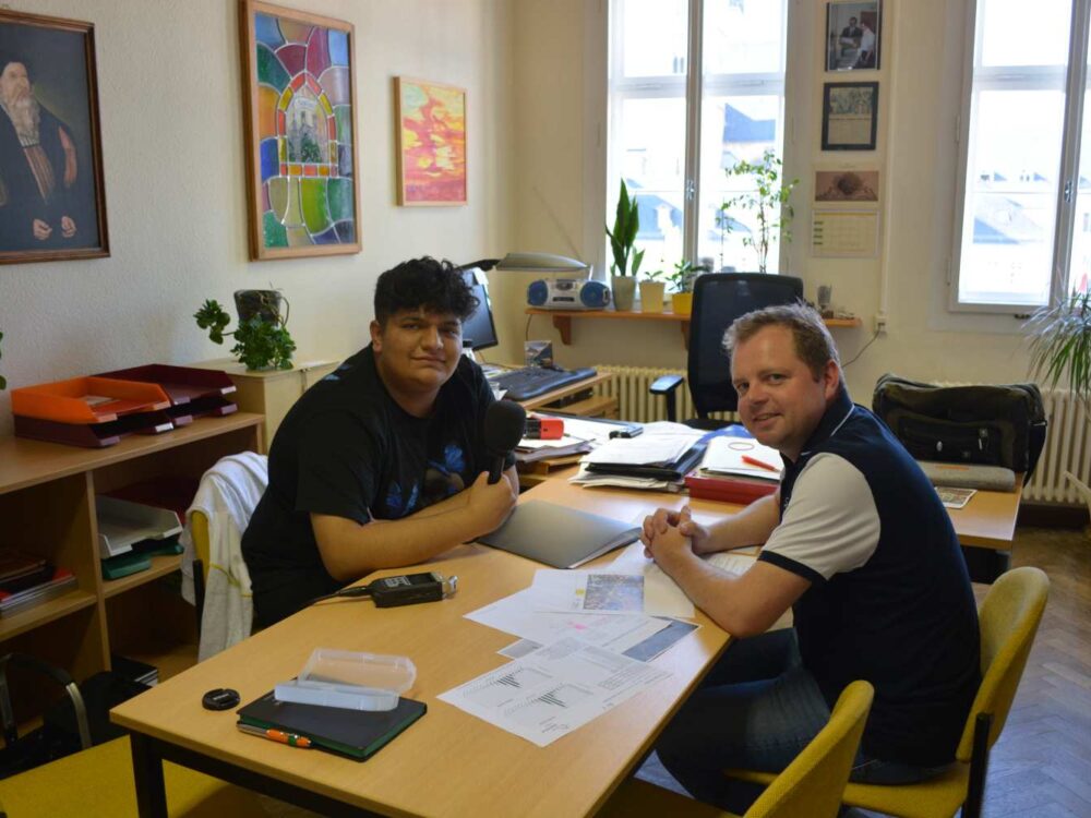 Ein Mann und ein Jugendlicher mit Migrationsgeschichte sitzen in einem Büro am Schreibtisch.