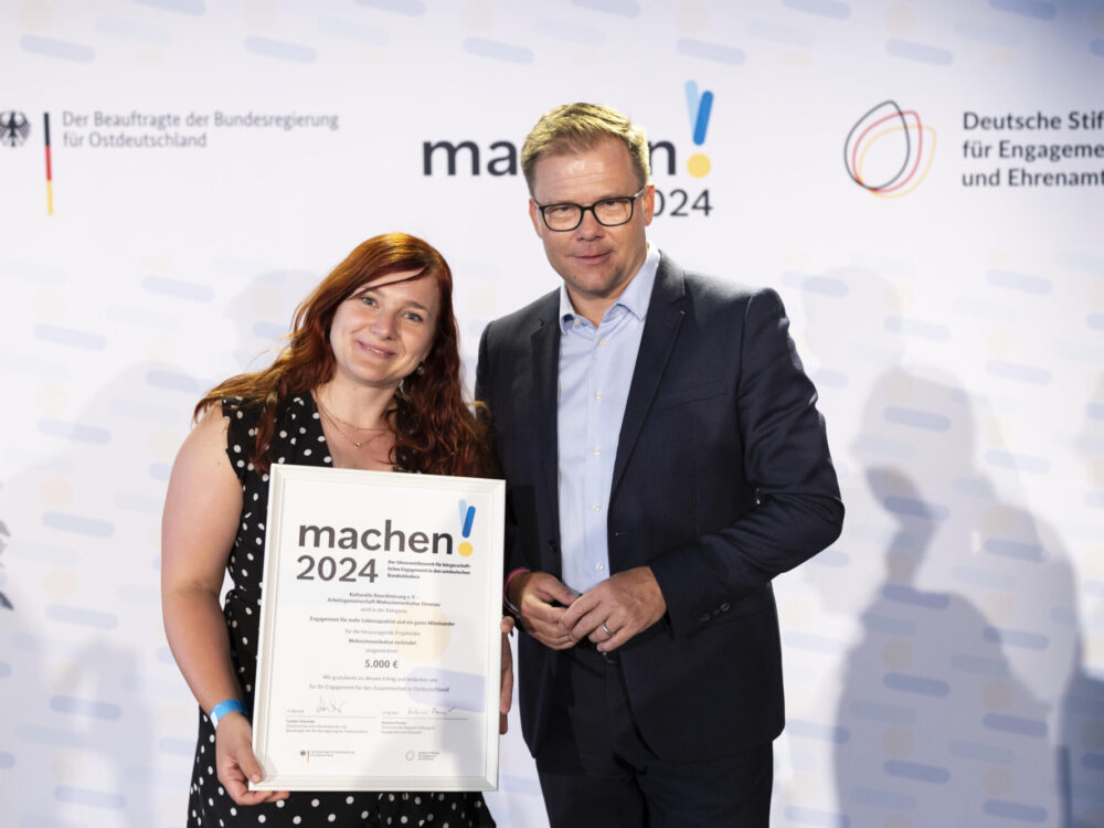 Eine Frau hält die Auszeichnung der Preisverleihung von „machen!2024“ in die Kamera und posiert neben Carsten Schneider.